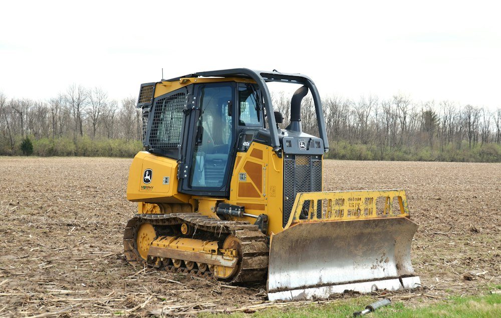 A compact track loader parked at the edge of a field beside a hardwood tree line